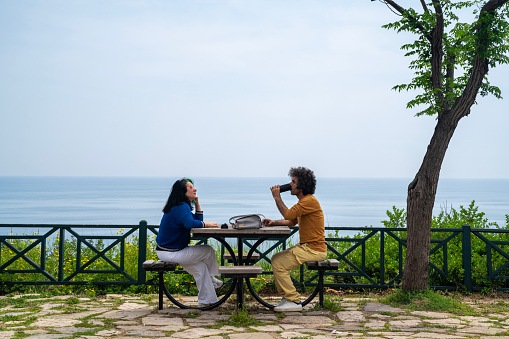full length photo of mature adult woman wearing a blue blouse and man wearing a yellow sweater sitting on park bench by sea. Tree is seen next to bench. Horizon over sea is seen. Shot under daylight.
