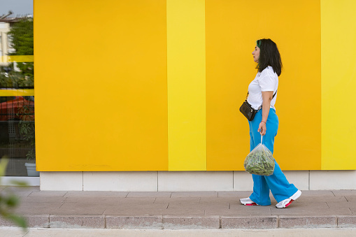 Full length photo of mature adult woman wearing blue pants and white t-shirt walking in front of yellow colored wall. She is carrying plastic bags. Shot under daylight.