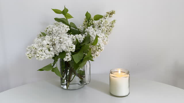 Burning candle in clear glass jar and white lilac bouquet on white table