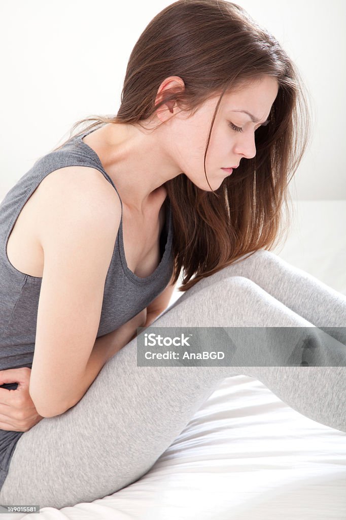 Young woman in gray clutching her stomach in pain Young woman in pain sitting on bed, on white background Abdomen Stock Photo