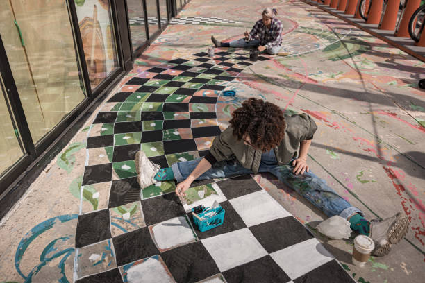 Two Female artists painting large sidewalk mural. They are dressed in casual work clothes. Exterior of public  sidewalk in downtown of large North American City.
