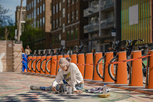 Young Caucasian woman artist painting sidewalk mural. She is dressed in casual work clothes. Exterior of public sidewalk in downtown of large North American City.