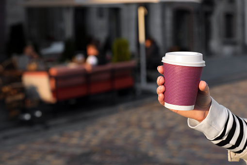 Woman holding paper takeaway cup on city street, closeup with space for text. Coffee to go