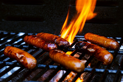 Bratwurst, hot dogs or polish sausage on a flaming charcoal grill.  One brat is being presented in a pair of tongs in front of the others.  All of them have beautiful appetizing grill marks as the flames kiss the meat sealing in the flavors.