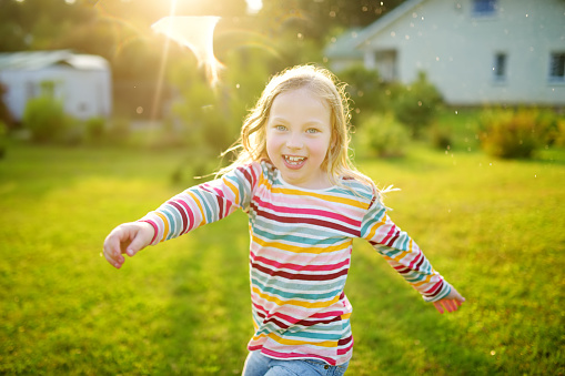 Adorable young girl playing with a sprinkler in a backyard on sunny summer day. Cute child having fun with water outdoors. Funny summer games for kids.
