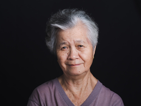 Portrait of an elderly Asian woman with short gray hair looking at the camera with a smile while standing on black background in the studio. Space for text. Aged people and health care concept