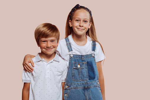 Two happy children hugging each other. Laughing teenage girl with her small brother having fun at studio, isolated over beige background. Children, childhood, family concept.