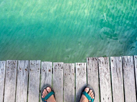 Top view of man feet wearing sandal standing on old wooden bridge over the clean and clear blue sea water near the beach in the island with copy space. Summer vacation holiday poster background.