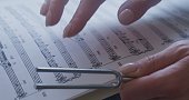 One woman's hand holds a tuning fork, the other traces the sheet music.