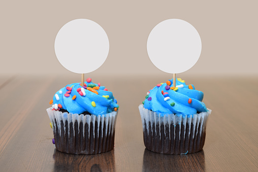 Two tempting blue frosted cupcakes resting on a modern brown wooding table with blank round cupcake toppers.