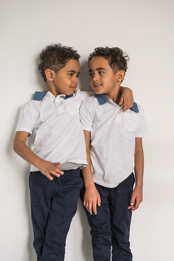 African American six year old twin boys looking at each other; one boy put his hand on his brother's shoulder; white wall in background