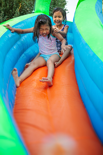 Happy asian girl sibling sliding down bouncy house water slide