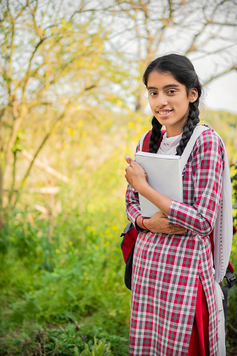 Outdoor image of happy rural Indian schoolgirl with a backpack holding a book and looking at the camera with a toothy smile.