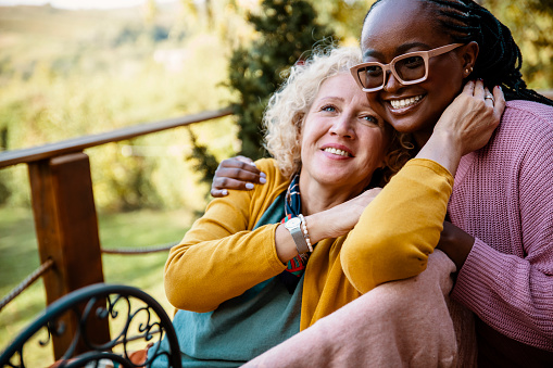 Mixed race female family relaxing in rehabilitation center in nature. Young African American Woman take care of disabled older Caucasian mother on a porch of nursing home. Copy space