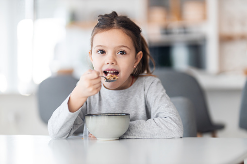 A cute little girl is sitting at the dining table at home and having a healthy morning meal, posing for the camera.