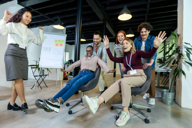 Young cheerful businesspeople in smart casual wear having fun while racing on office chairs and smiling