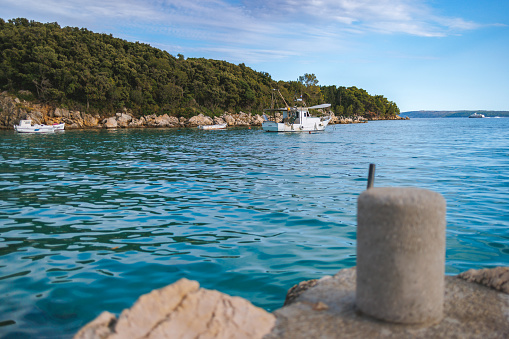 View on fishing boats in the bay near coastline going to the open sea under the blue sky on summer day, concrete bollard on pier cast on rocky coast, small jetty for mooring