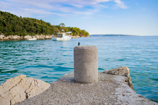 Cylinder shaped concrete bollard on pier cast on rocky coast, small jetty for mooring fishing boats, view from bay on coastline and open sea under the blue sky in summer