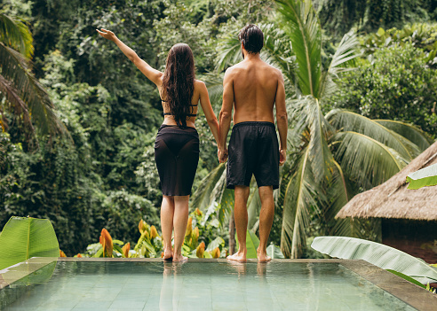 Rear view shot of young couple standing on the edge of the pool holding hands and looking at a view. Man and woman at luxury holiday resort.