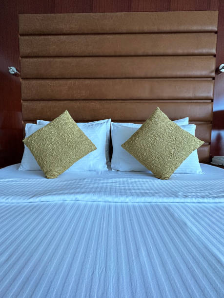 istock Full frame image of stack of white hotel pillows leaning on leatherette headboard, white duvet bed cover, gold cushions, focus on foreground 1490281309