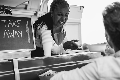 African senior woman serving take away food inside food truck - Soft focus on chef face - Black and white editing