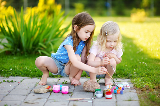 Two cute little sisters painting on stones together on hot summer day
