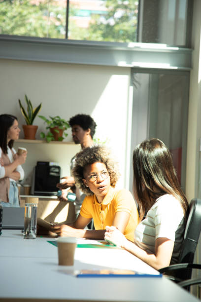 istock Two female coworkers share ideas while two colleagues have coffee break in the background. Vertical image. Copy space. 1490293230