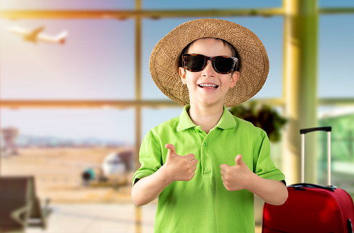 Happy child with hat and sunglasses holding thumbs up at airport.