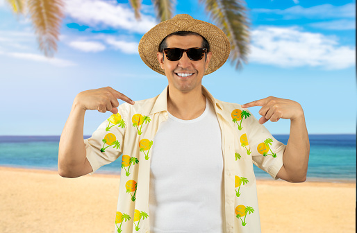 Latin man on vacation wearing floral shirt hat sunglasses at tropical beach looking confident with smile on face, pointing oneself with fingers proud and happy.