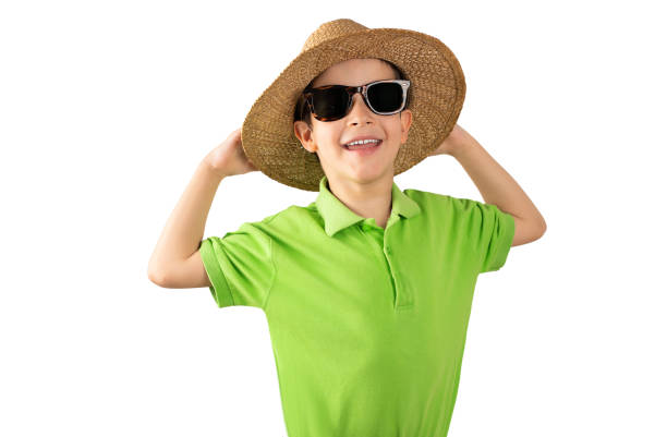 Child on vacation wearing green t-shirt hat and sunglasses over white isolated background picking up the hat.Kid with happy face smiling and looking at the camera. Positive person