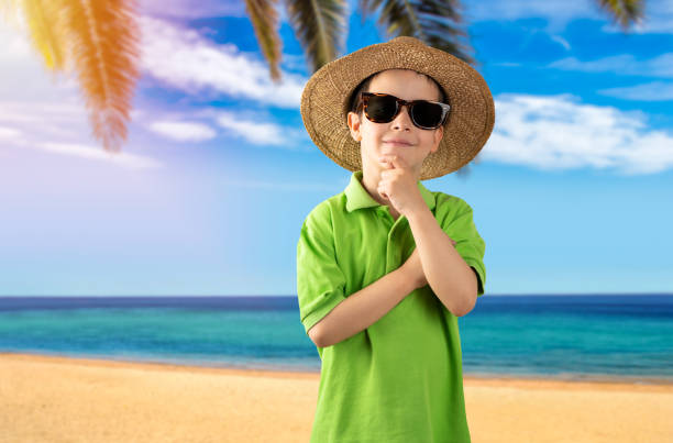 Child on vacation wearing sunglasses wearing green t-shirt hat tropical beach with hand on chin thinking about question, pensive expression. pensive face. Concept of doubt and look aside