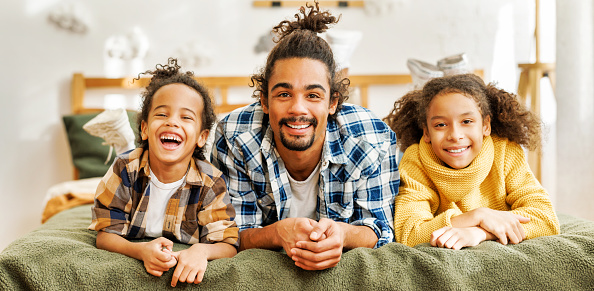 Happy afro american family father and cute kids son and daughter relaxing at home in bed, overjoyed excited children spending time with dad