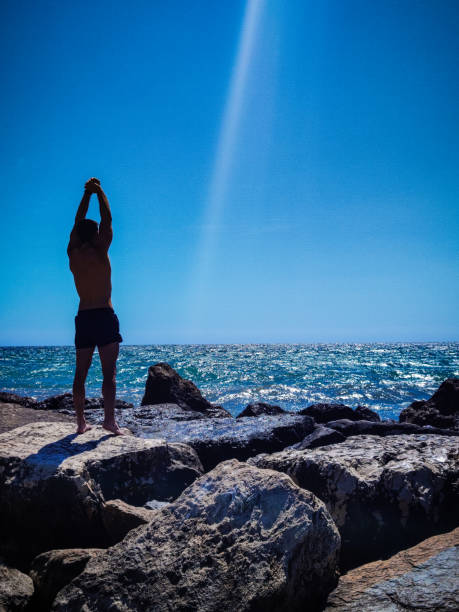 Man from behind stretching on top of the rocks of the beach in front of the sea. Dark silhouette strong man without shirt before doing sport outdoors. Copy space. Illuminated scene with a sunbeam.