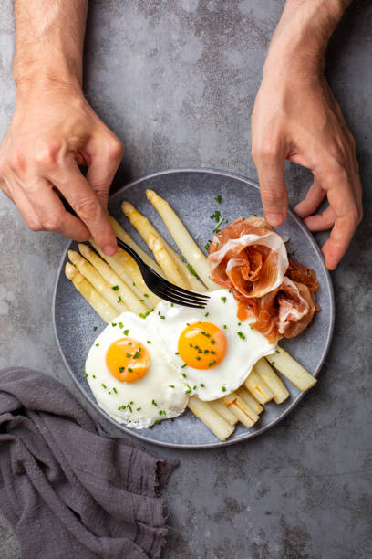 Eating white asparagus with fried eggs, crudo prosciutto or  ham, spring onion and butter dressing. Mans hands holding a fork. Top view, dark grey background. stock photo