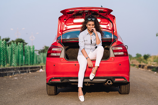 Outdoor image of a young girl having fun on a road trip in car, She came out of the car back trunk and enjoying the nature with smile and joy. Concept of weekend vacation and long road trip.