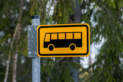 Yellow local bus stop sign in Finland, with green pine trees in the background.