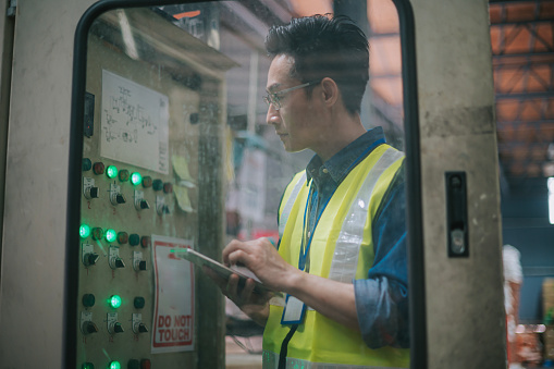 Asian Chinese male maintenance engineer checking on factory control panel with digital tablet
