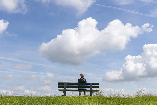 Texel, The Netherlands-May 2022; Low angle view of a woman sitting on a park bench on top of a dike looking in invisible distance with large cumulus clouds above in otherwise blue sky