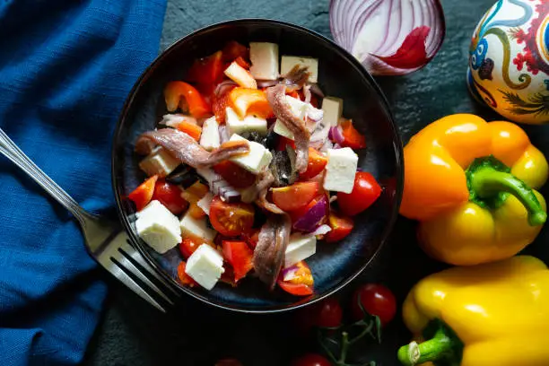 Greek salad of tomatoes, peppers, red onion, anchovies, black olives and feta cheese in a black bowl on a slate background