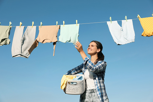 Smiling woman holding basket with baby clothes near washing line for drying against blue sky outdoors