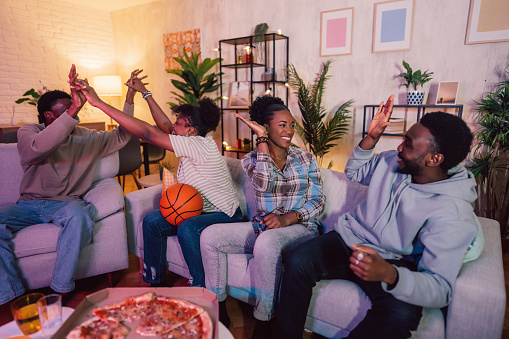 Beautiful black family sitting together, eating pizza and watching a basketball game in the living room at night.