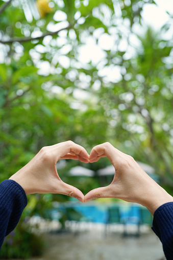 The cropped shot highlights a woman's hand raised, forming a heart shape against the backdrop of a stunning tropical greenery resort. This image represents the harmonious beauty of nature and conveys a sense of relaxation and a perfect holiday vacation destination.