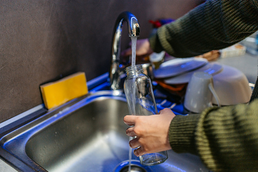 Close-up of a young woman refiling her glass water bottle with water in the domestic kitchen.
