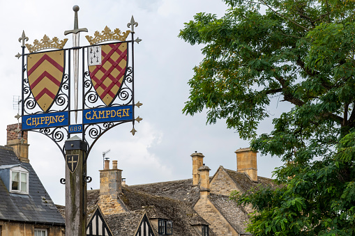 Chipping Campden, UK-August 2022; Low angle view of the town shields of wrought iron with a sword on a wooden post at the start of the Cotswold Way, with roofs of limestone cottages in background