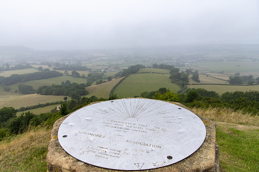 Dursley, UK-August 2022; Close up view from the Cotswold Edge on Coaley Peak of the topographic steel plate on top the trig point along the Cotswolds Way with panoramic view over the hilly landscape