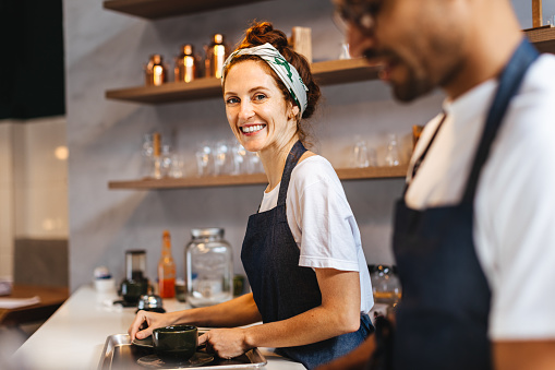 Caucasian female barista smiling at the camera while working alongside her colleague in a coffee shop. Cafe workers wearing aprons and preparing hot drinks for their customers.