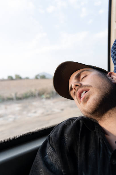 A young Caucasian man wearing a cap is taking a nap during a bus trip with the head resting on the window