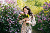 Young woman walks in garden and enjoys by lilac bouquet in her hands .