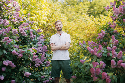 Young smiling man walks in garden and enjoys by blooming lilac shrubs.