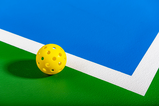 A low angle view of a yellow Pickleball ball sitting on the corner of a green and blue Pickleball court.